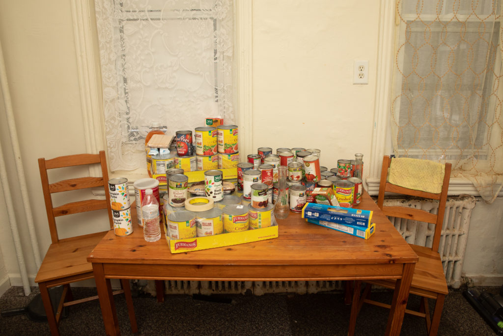 a wooden table sits between to wooden chairs in a room. The table top is full of canned goods.